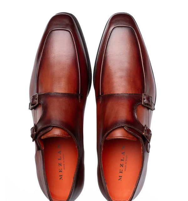 A pair of men's double monk strap loafers in a rich cognac brown leather, shown from a top-down perspective on a white background. The brand name 'MEZLAN' is printed in black on the orange interior lining.