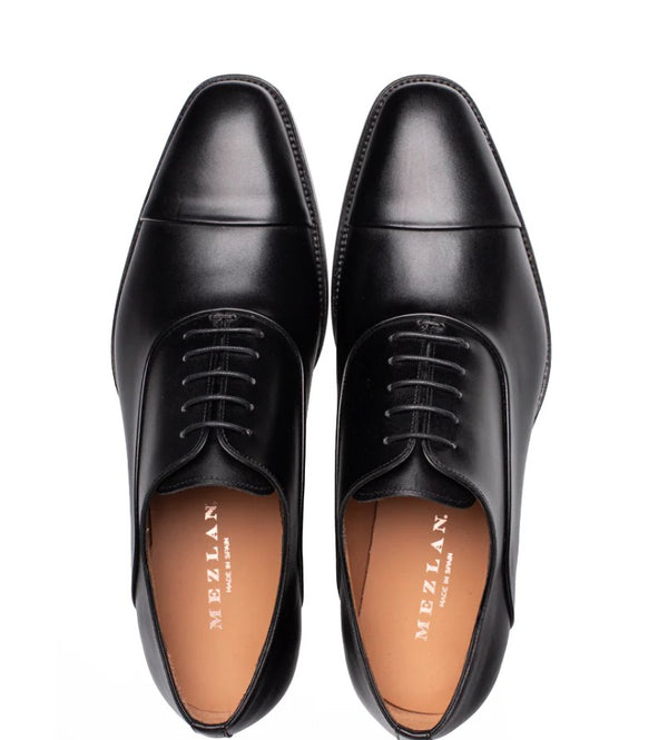 A pair of black leather dress shoes with a cap toe and laces, shown from a top-down view on a white background. The tan insole displays the brand name 'MEZLAN'.