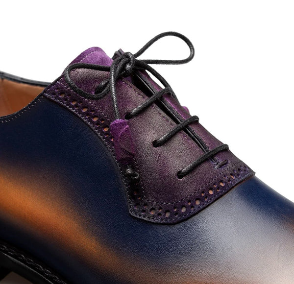 A close-up of a men's lace-up dress shoe showing a glossy dark blue leather toe, a purple suede overlay, and black laces on a white background.