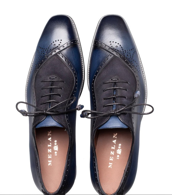 A pair of men's wingtip oxford shoes in navy blue and black brogue leather, shown from a top-down view on a white background. The brand name 'MEZLAN' is visible on the insoles.