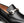 A close-up of a black leather loafer featuring a prominent silver-tone metal horsebit detail across the vamp, set against a clean white background.