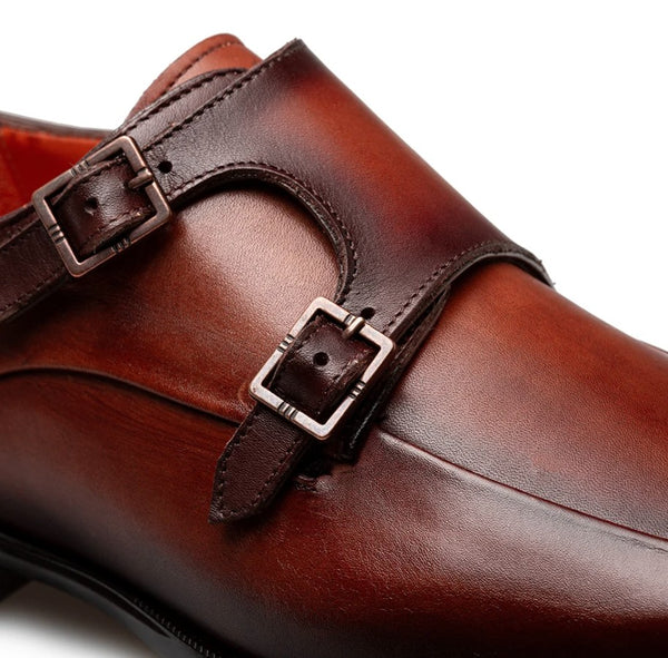 A close-up of a brown leather double monk strap shoe with two dark brown straps and silver-toned buckles, shown on a white background.