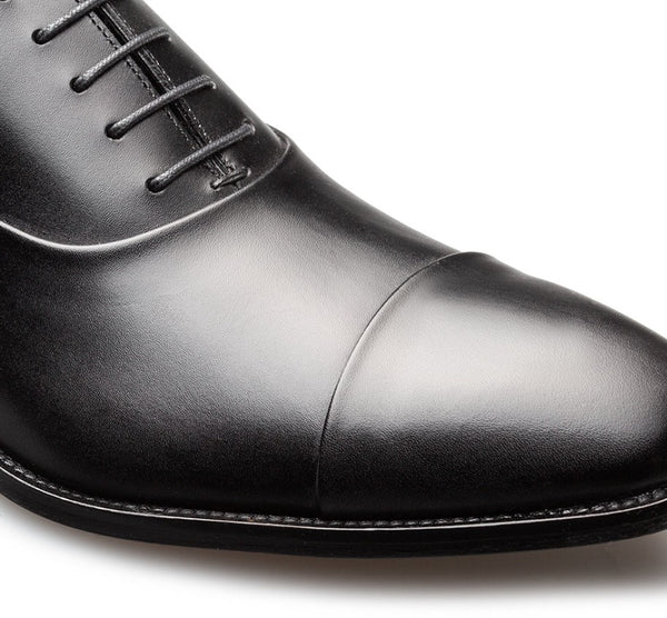 Close-up of a black leather oxford dress shoe showing the cap toe, laces, and side stitching against a white background.