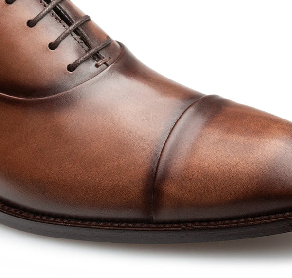 Close-up of a brown leather cap-toe oxford shoe showing the toe, side panel, laces, and stitching on a white background.