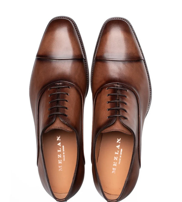 A pair of brown leather dress shoes with a cap toe and laces, shown from a top-down view on a white background. The brand name 'MEZLAN' is visible on the insoles.
