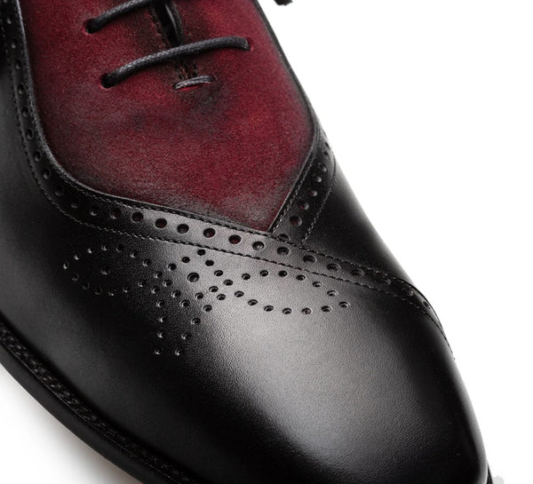 A close-up of a men's wingtip oxford shoe with a black brogue leather toe and black laces, transitioning into a deep burgundy red suede vamp on a white background.