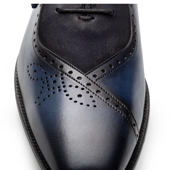 A close-up of a men's wingtip dress shoe in dark navy blue, showing the glossy leather toe, perforated brogue detailing, and dark suede vamp against a white background.