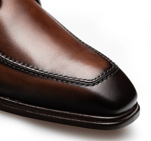 Close-up of a brown leather dress shoe toe cap showing the smooth vamp, decorative stitching, and dark brown sole edge on a white background.
