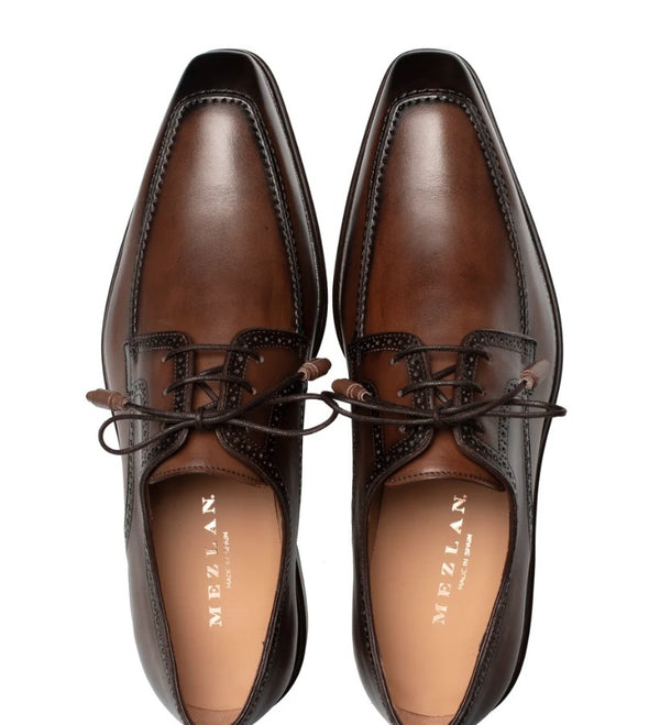A pair of brown leather dress shoes with a glossy finish, featuring a cap toe, decorative brogue detailing, and dark brown laces. The brand name 'MEZLAN' is printed in gold on the tan insole.