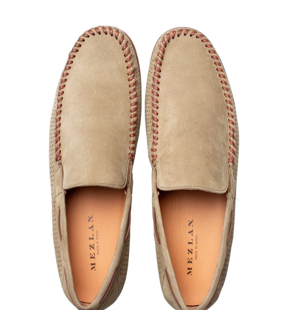 A pair of beige suede slip-on shoes with brown contrast stitching, shown from a top-down view on a white background. The brand name 'MEZLAN' is visible on the insole.